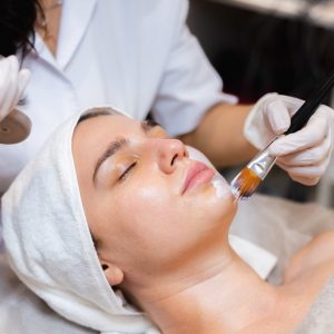 Beautician with a brush applies a white moisturizing mask to the face of a young girl client in a spa beauty salon