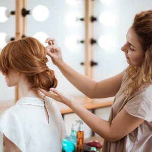 Female hairdresser making hairstyle to beautiful redhead girl in beauty salon. Copy space.